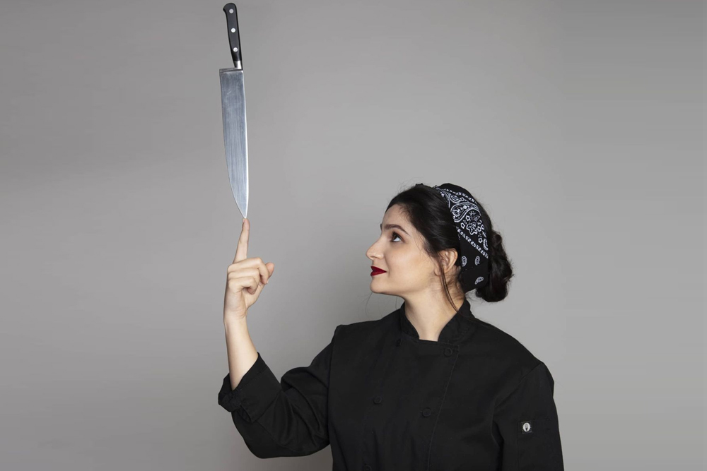 Chef Tara Khattar in a black chef's jacket, balancing a chef's knife on her fingertip
